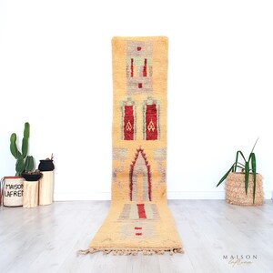 May include: A long, rectangular rug with a cream-colored background and geometric patterns in red, gray, and cream. The rug features a central design with a series of rectangular and arched shapes. The rug is made of a textured material and has a fringe at the bottom.