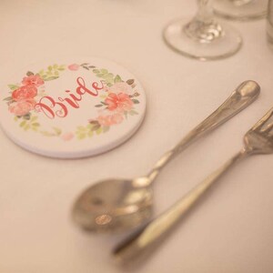 May include: A white round coaster with a floral design and the word "Bride" in red lettering.