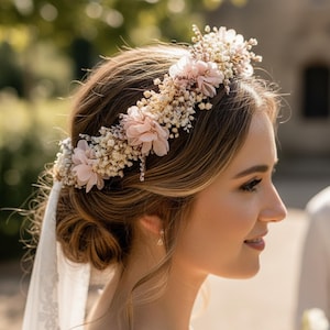 Puede incluir: Una mujer con una corona floral con flores rosas y blancas. La corona está sobre su cabello castaño y lleva un vestido de encaje blanco. Un velo blanco está sujeto a la corona.