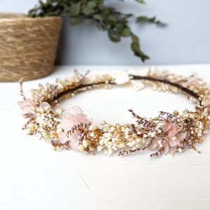 Puede incluir: Una corona floral con base marrón adornada con flores blancas y beige secas, y flores de tela rosa pálido. La corona tiene una estética delicada y natural, ideal para ocasiones especiales.
