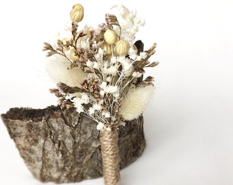 Puede incluir: Un pequeño prendedor de flores secas con una mezcla de flores blancas, crema y marrones y cápsulas de semillas. Los tallos están envueltos con hilo. El prendedor se exhibe sobre un trozo de madera.