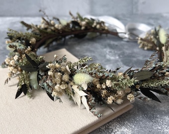 May include: A dried flower crown with white, green, and brown flowers and leaves. The crown is on a white surface.