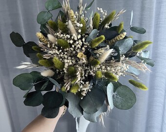 Pode incluir: Um buquê de flores secas com tons de verde, branco e bege. O arranjo inclui folhas de eucalipto, gramíneas verdes fofas e pequenas flores brancas. O buquê é amarrado com uma fita branca.