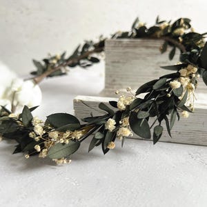 May include: A handcrafted floral crown featuring dark green eucalyptus leaves and small, delicate white flowers. The crown is arranged on a light-colored wooden surface, creating a natural and elegant aesthetic. Perfect for weddings or special occasions.
