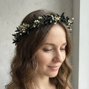 May include: A dark green floral crown with small white flowers and dark green leaves. The crown is worn on a person's head. The flowers and leaves are arranged in a circular pattern, creating a natural and elegant look. Perfect for weddings or special occasions.