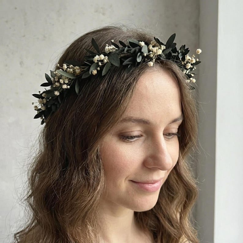 May include: A dark green floral crown with small white flowers and dark green leaves. The crown is worn on a person's head. The flowers and leaves are arranged in a circular pattern, creating a natural and elegant look. Perfect for weddings or special occasions.