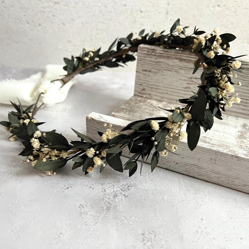 May include: A handcrafted floral crown featuring dark green leaves and small, cream-colored flowers. The crown is tied with a white ribbon. This botanical headpiece is suitable for weddings, festivals, or special occasions.