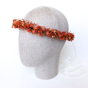 May include: A floral crown made of dried orange flowers and greenery. The crown is on a mannequin head and has a white ribbon attached.