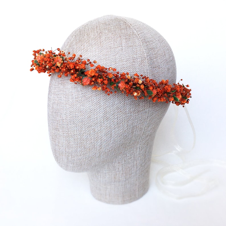 May include: A floral crown made of dried orange flowers and greenery. The crown is on a mannequin head and has a white ribbon attached.
