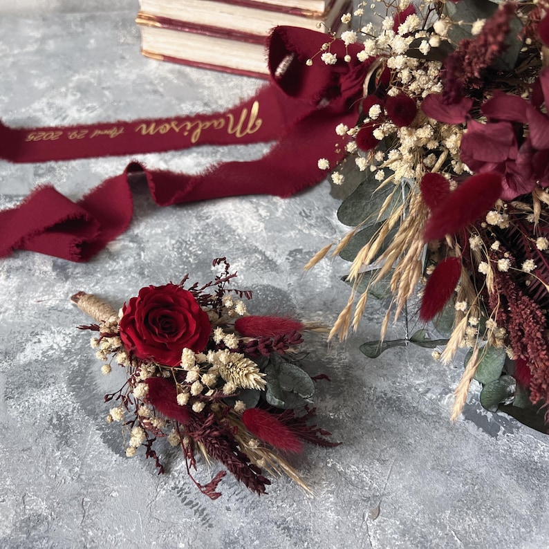 May include: A burgundy floral arrangement featuring a red rose, dried flowers, and greenery. A matching boutonniere is also present. A burgundy ribbon with gold text that reads "Madison and 29, 2025" is also visible. The arrangement is set against a gray background.