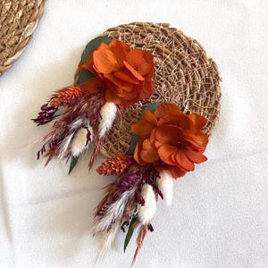 Könnte beinhalten: Ein Paar orangefarbene getrocknete Blumenohrringe mit weißen und burgunderroten getrockneten Gräsern. Die Ohrringe werden auf einem geflochtenen Strohhintergrund präsentiert.