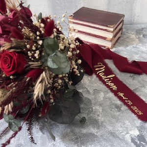 May include: A burgundy bouquet featuring a red rose, dried flowers, and greenery. A burgundy ribbon with the name "Madison" and the date "April 29, 2025" is draped near the bouquet. A stack of burgundy books is in the background.