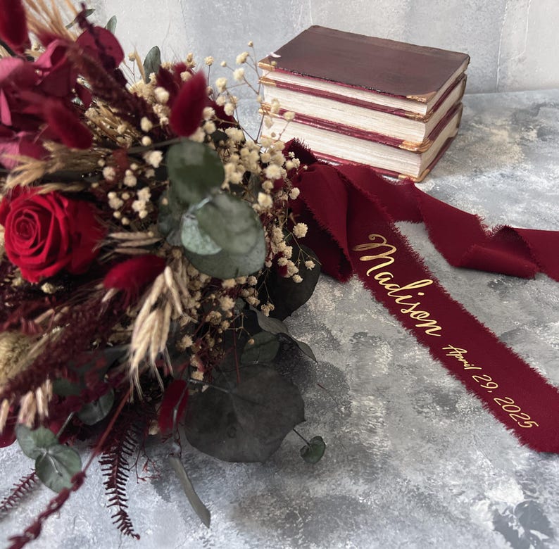 May include: A burgundy bouquet featuring a red rose, dried flowers, and greenery. A burgundy ribbon with the name "Madison" and the date "April 29, 2025" is draped near the bouquet. A stack of burgundy books is in the background.
