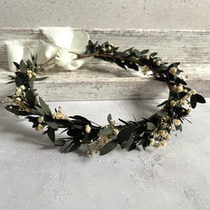 May include: A handcrafted floral crown featuring dark green leaves and small white flowers. The crown is tied with a white ribbon. This botanical headpiece is suitable for weddings, festivals, or special occasions.