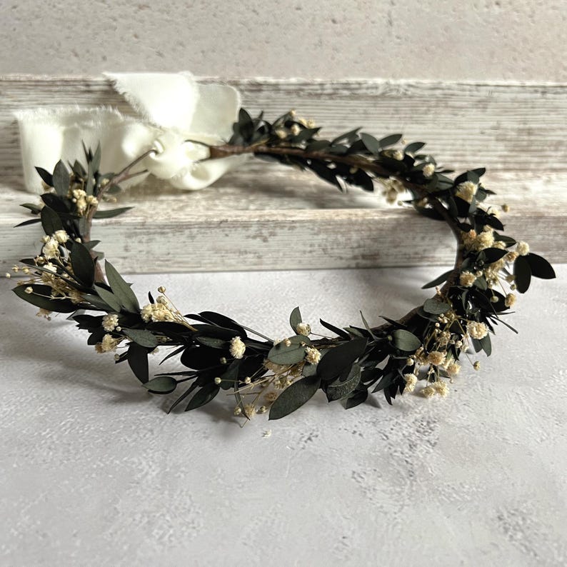 May include: A handcrafted floral crown featuring dark green leaves and small white flowers. The crown is tied with a white ribbon. This botanical headpiece is suitable for weddings, festivals, or special occasions.