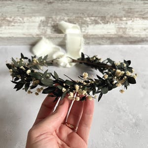 May include: A handcrafted floral crown featuring dark green leaves and small white flowers. The crown is held by a hand, with a cream-colored ribbon attached. The crown is designed for special occasions.