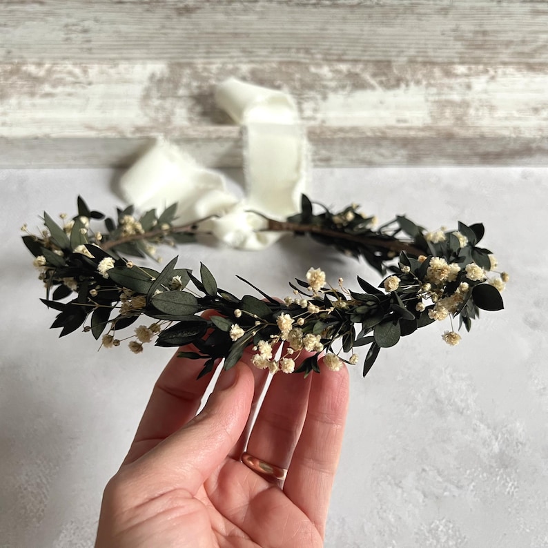 May include: A handcrafted floral crown featuring dark green leaves and small white flowers. The crown is held by a hand, with a cream-colored ribbon attached. The crown is designed for special occasions.