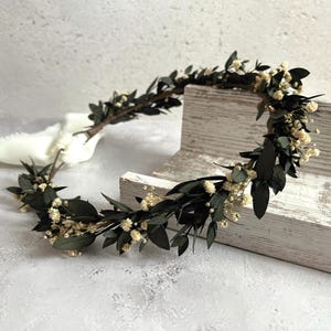 May include: A handcrafted floral crown featuring dark green leaves and small, delicate white flowers. The crown is tied with a white ribbon. The natural elements create a rustic and elegant aesthetic, perfect for weddings or special occasions.
