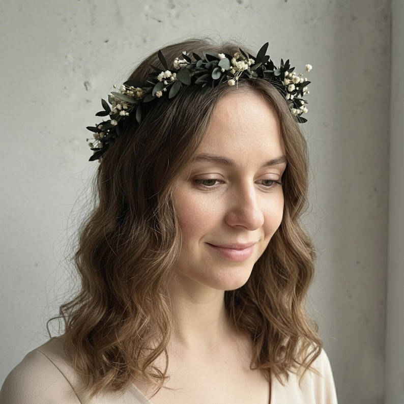 May include: A dark green floral crown with small white flowers and green leaves. The crown is worn on a person's head. The person has long, wavy brown hair and is wearing a light-colored top.