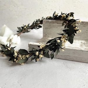 May include: A handcrafted floral crown featuring dark green leaves and small, delicate white flowers. The crown is arranged in a circular shape, perfect for weddings or special occasions. The natural elements create a rustic and elegant aesthetic.