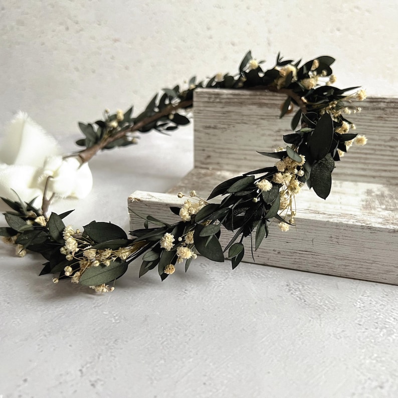 May include: A handcrafted floral crown featuring dark green leaves and small, delicate white flowers. The crown is arranged in a circular shape, perfect for weddings or special occasions. The natural elements create a rustic and elegant aesthetic.