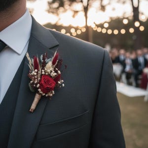 May include: A dark gray suit with a red rose boutonniere. The boutonniere features a red rose, dried flowers, and greenery, wrapped with twine. The suit has a white dress shirt and a dark green tie. The background is blurred with soft lighting.