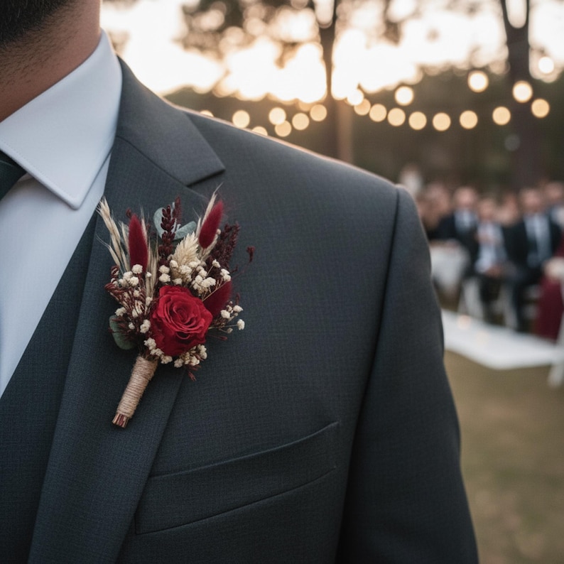May include: A dark gray suit with a red rose boutonniere. The boutonniere features a red rose, dried flowers, and greenery, wrapped with twine. The suit has a white dress shirt and a dark green tie. The background is blurred with soft lighting.