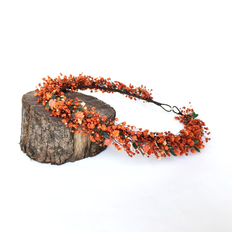May include: A floral crown made of dried orange flowers and greenery. The crown is on a wooden log and is ready to be worn.