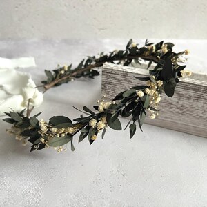 May include: A handcrafted floral crown featuring dark green leaves and small, cream-colored flowers. The crown is made of dried greenery and is arranged in a circular shape. The crown is a natural, rustic accessory.