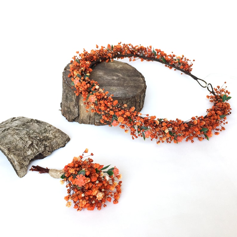 May include: A set of two orange flower crowns, one large and one small. The larger crown is on a wooden log, and the smaller crown is on a white surface. Both crowns are made of dried flowers and have a wire base.
