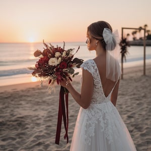 May include: A bride stands on a beach at sunset, holding a bouquet of red and cream-colored flowers with burgundy ribbons. She wears a white lace wedding dress and a veil with a bow in her hair. The ocean and a wooden arch are in the background.