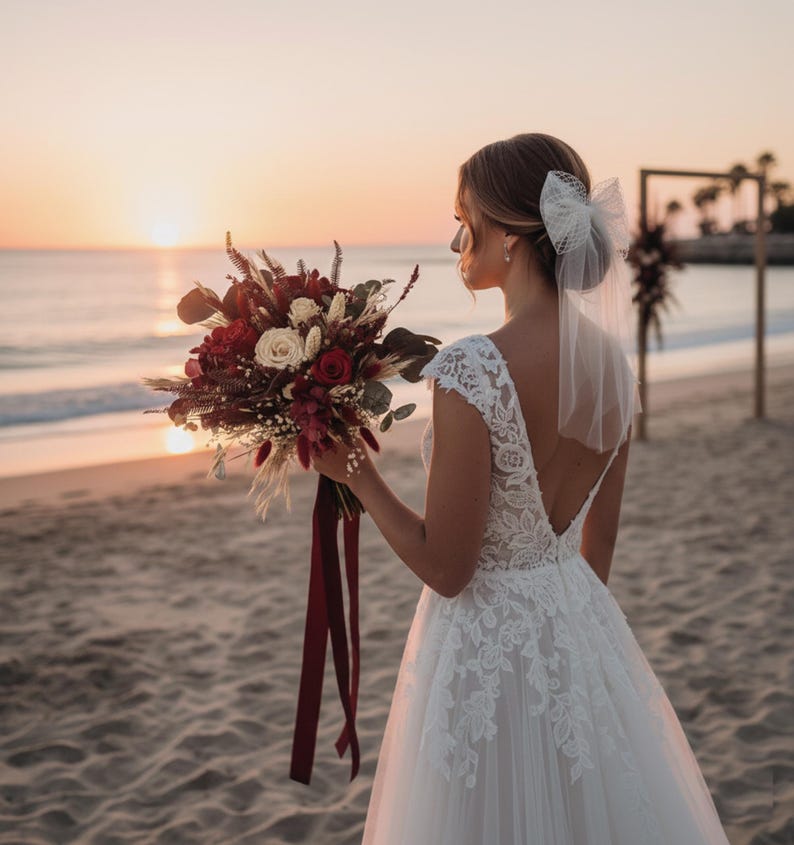 May include: A bride stands on a beach at sunset, holding a bouquet of red and cream-colored flowers with burgundy ribbons. She wears a white lace wedding dress and a veil with a bow in her hair. The ocean and a wooden arch are in the background.