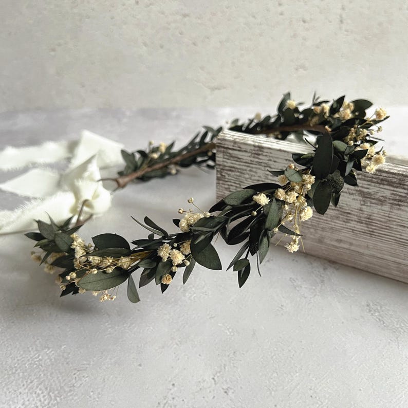 May include: A handcrafted floral crown featuring dark green leaves and small, delicate white flowers. The crown is arranged on a wooden box and includes a white ribbon, perfect for weddings or special occasions.