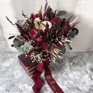 May include: A bridal bouquet featuring red roses, burgundy accents, and cream-colored flowers. The bouquet includes dried wheat, eucalyptus, and baby's breath. A burgundy ribbon with gold script reading "Madison" is tied around the stems.
