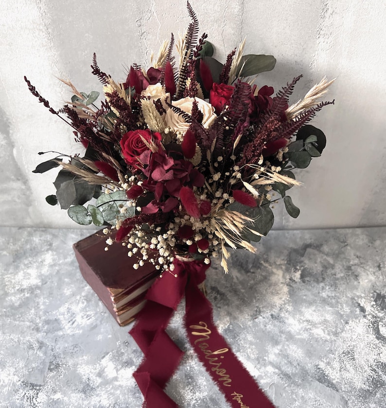 May include: A bridal bouquet featuring red roses, burgundy accents, and cream-colored flowers. The bouquet includes dried wheat, eucalyptus, and baby's breath. A burgundy ribbon with gold script reading "Madison" is tied around the stems.