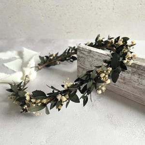 May include: A handcrafted floral crown featuring dark green leaves and small, delicate white flowers. The crown is made of natural materials and is designed to be worn on the head. Perfect for weddings, festivals, or special occasions.