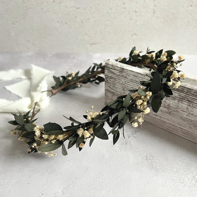May include: A handcrafted floral crown featuring dark green leaves and small, delicate white flowers. The crown is made of natural materials and is designed to be worn on the head. Perfect for weddings, festivals, or special occasions.