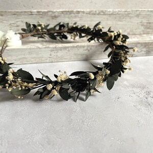 May include: A handcrafted floral crown featuring dark green leaves and small, delicate white flowers. The crown is arranged in a circular shape, perfect for weddings or special occasions. The crown is set against a neutral background.