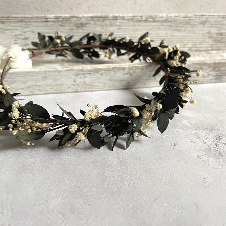 May include: A handcrafted floral crown featuring dark green leaves and small, delicate white flowers. The crown is arranged in a circular shape, perfect for weddings or special occasions. The crown is set against a neutral background.