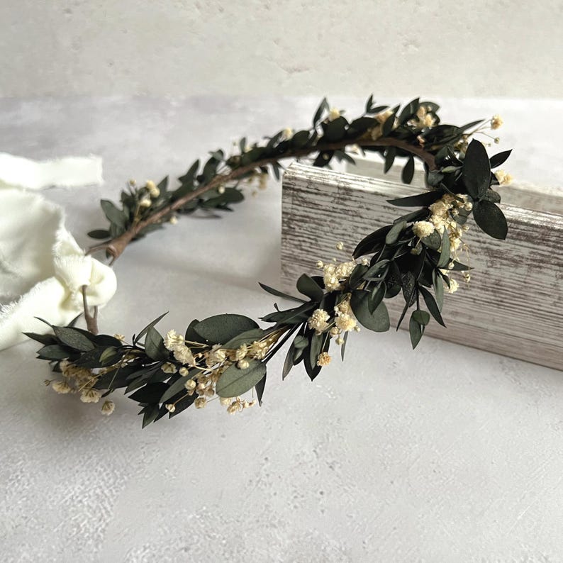 May include: A handcrafted floral crown featuring dark green leaves and small white flowers. The crown is made with a natural, rustic aesthetic, perfect for weddings or special occasions. The crown is approximately 12 inches in diameter.