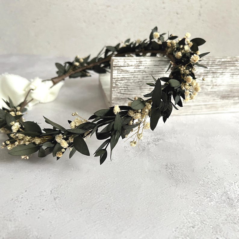 May include: A handcrafted floral crown featuring dark green leaves and small, cream-colored flowers. The crown is tied with a white ribbon. The foliage is arranged around a brown stem, creating a natural and elegant design, perfect for weddings or special occasions.