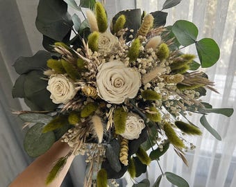 Pode incluir: Um buquê de noiva com rosas marfim, folhas de eucalipto verdes e várias ervas secas. O arranjo inclui detalhes verdes e bege fofos, criando um visual natural e texturizado. O buquê é segurado contra um fundo macio e neutro.