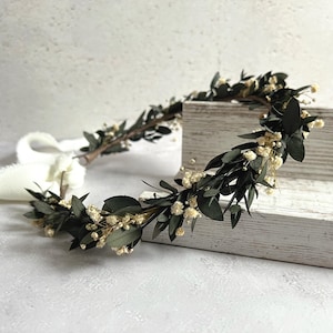 May include: A handcrafted floral crown featuring dark green leaves and small, delicate white flowers. The crown is arranged on a wooden surface, with a white ribbon attached. This botanical headpiece is suitable for weddings or special occasions.