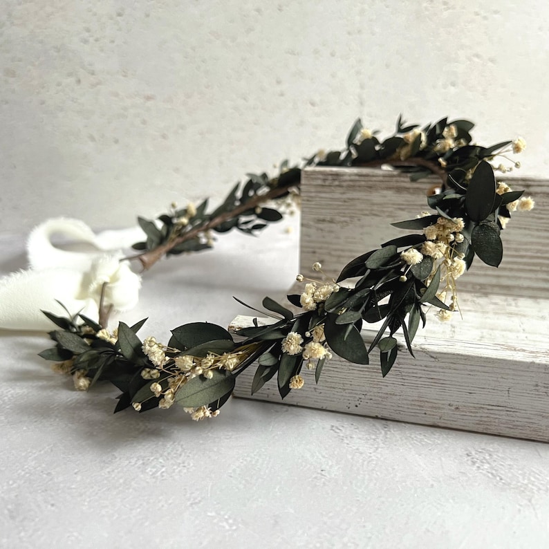 May include: A handcrafted floral crown featuring dark green leaves and small, delicate white flowers. The crown is arranged on a wooden surface, with a white ribbon attached. This botanical headpiece is suitable for weddings or special occasions.