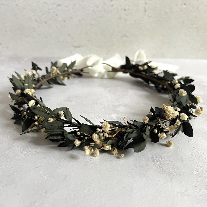 May include: A circular floral crown featuring dark green leaves and small, cream-colored flowers. The crown is tied with a white ribbon. This handcrafted headpiece is suitable for weddings, festivals, or special occasions.