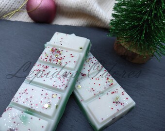 May include: Two white wax melts with a green border and red and gold glitter. The wax melts are stacked on top of each other. The background is a dark gray surface.