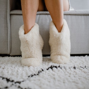 Op de afbeelding: Een paar witte, pluche pantoffels met een zachte, donzige textuur. De pantoffels worden gedragen door iemand die op een bank zit.