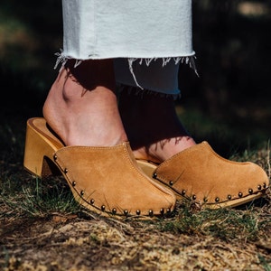 May include: A pair of brown suede clogs with wooden heels and metal studs. The clogs are worn by a person standing on grass.