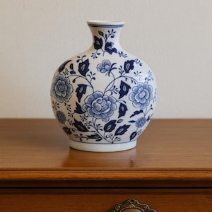 Pode incluir: Um vaso de cerâmica branco com corpo arredondado e pescoço estreito. O vaso é decorado com um padrão floral azul. O vaso está sobre uma superfície de madeira.