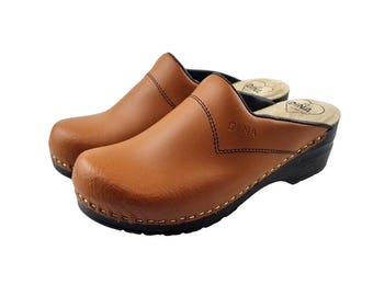 May include: A pair of brown leather clogs with a black sole and a rounded toe. The clogs have a stitched design and the word "DINA" is visible on the side. The interior has a heart-shaped logo.